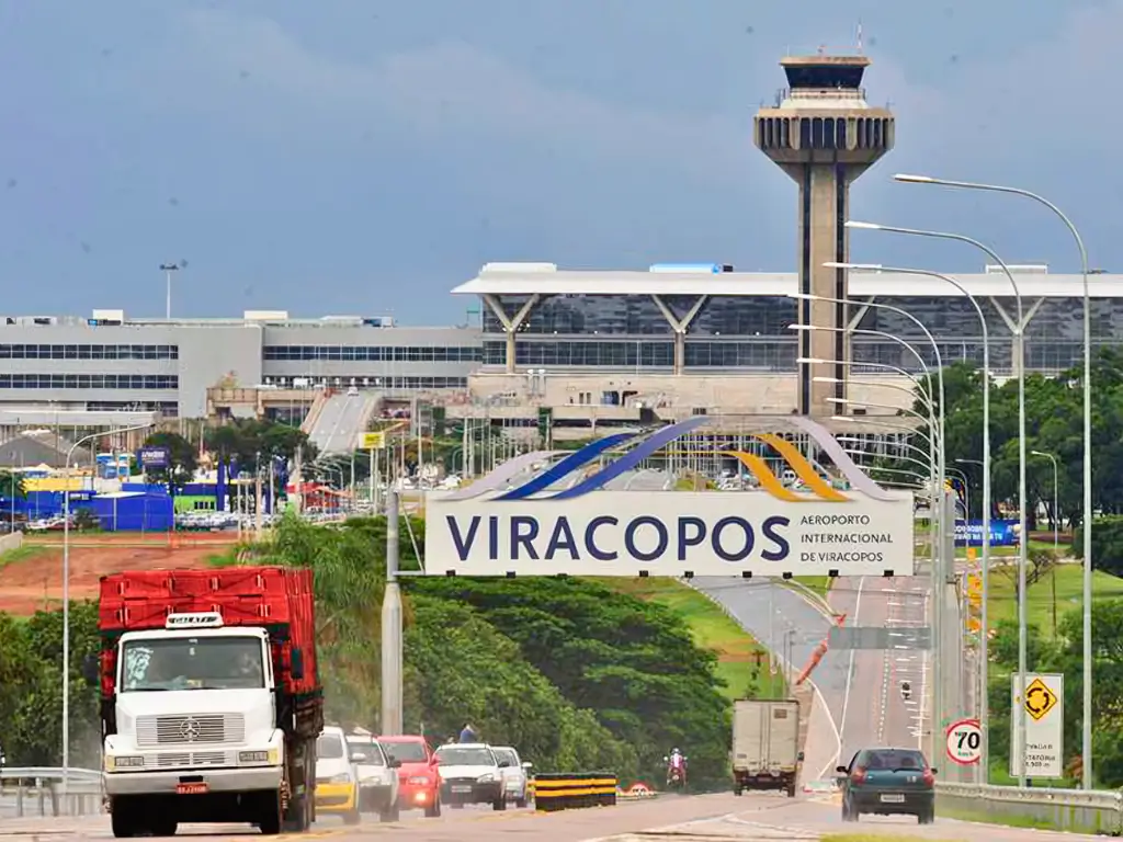 Vista da entrada do Aeroporto Internacional de Viracopos com um portal grande com o nome do aeroporto, tráfego de veículos na rodovia em primeiro plano, e a torre de controle e terminais ao fundo em um dia nublado.