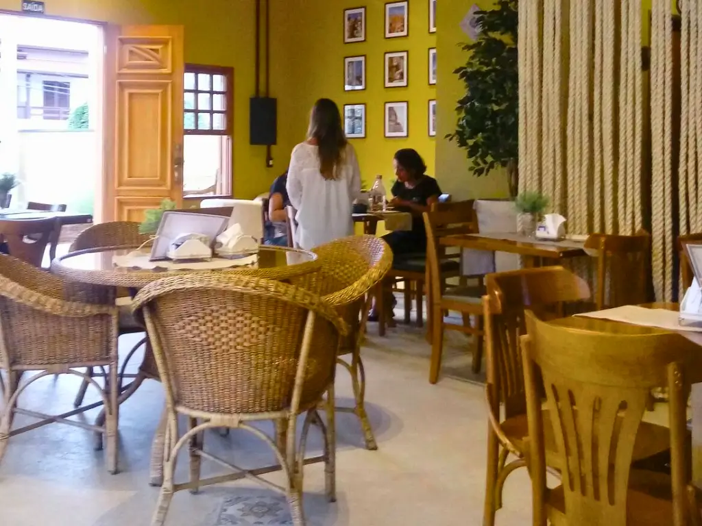 Interior de uma cafeteria com paredes amarelas decoradas com quadros variados, mesas de vime e clientes desfrutando do ambiente relaxante.