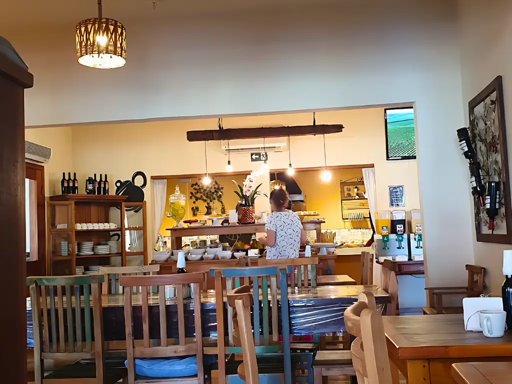 Interior do Casarão Café Colonial com decoração rústica, mesas de madeira, buffet de café da manhã e luminária pendente, criando um ambiente acolhedor e familiar.