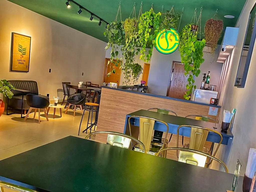 Interior de uma cafeteria moderna e aconchegante com plantas suspensas, decoração verde e amarela, mesas e cadeiras metálicas, e uma atmosfera relaxante com assentos confortáveis.