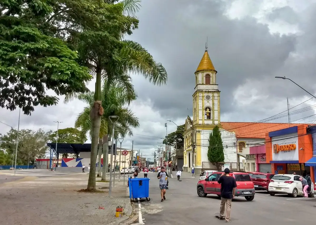 Rua de uma cidade com uma igreja de torre amarela ao fundo, pessoas caminhando e carros estacionados, com árvores e lojas ao redor. Cotia-SP