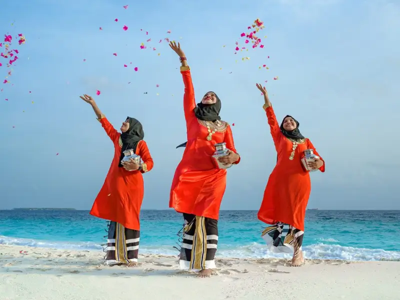 Três mulheres maldivas em trajes tradicionais vermelhos e dourados jogam pétalas de flores no ar na praia. Elas usam lenços na cabeça e sorriem enquanto celebram, com o mar azul ao fundo.