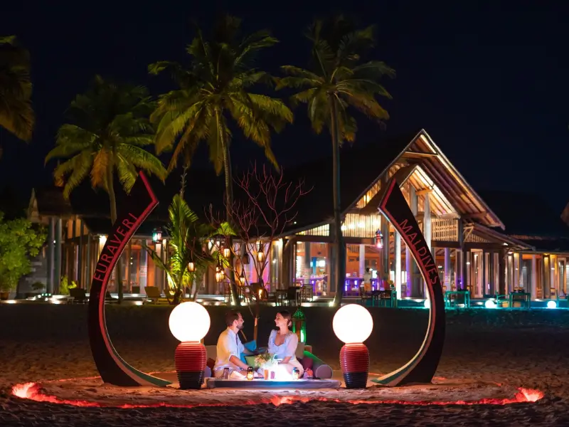 Casal jantando à luz de velas na praia em um resort nas Maldivas à noite, sob palmeiras. Eles estão sentados em almofadas vermelhas ao redor de uma mesa baixa decorada, com um grande enfeite escrito 'Maldives' ao fundo e construções iluminadas.