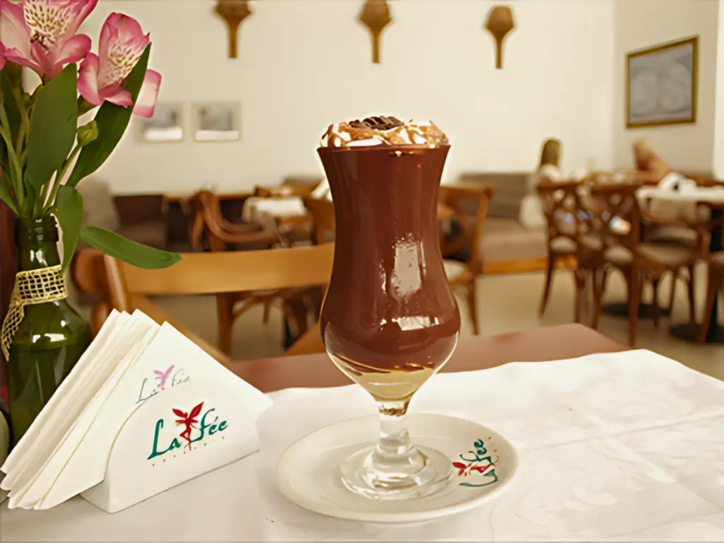 Um delicioso copo de café gelado coberto com chantilly e calda de chocolate, servido em uma mesa com guardanapos personalizados do café La Fée, em um ambiente elegante e tranquilo de cafeteria, com mesas de madeira ao fundo.