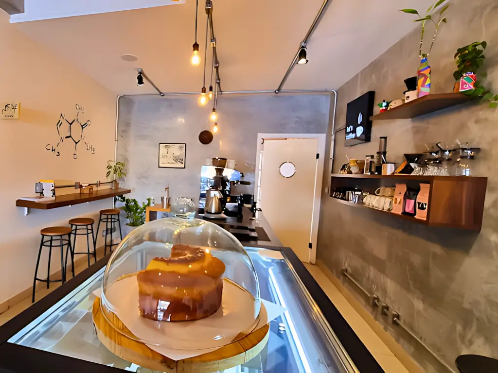 Interior minimalista de uma cafeteria moderna com um bolo sob uma cúpula de vidro em primeiro plano, máquinas de café e prateleiras com produtos decorativos ao fundo, em um espaço decorado com iluminação pendente e paredes de concreto.