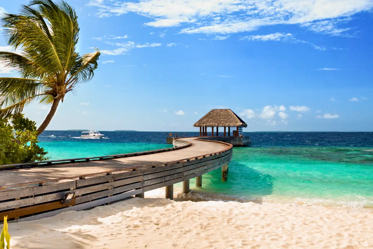 Praia paradisíaca com areia branca, mar azul-turquesa, um píer de madeira levando a um gazebo coberto de palha sobre a água e um barco ao fundo sob um céu azul com nuvens.