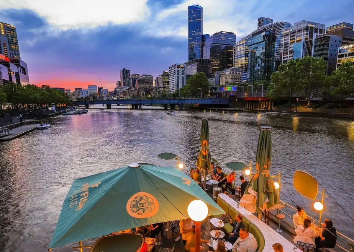 Pessoas desfrutando de uma refeição ao ar livre em um restaurante à beira do rio em Melbourne ao entardecer, com o horizonte da cidade ao fundo.