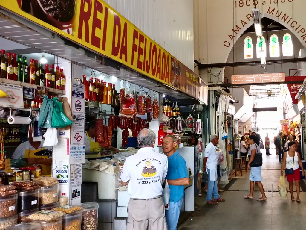 Interior do Mercado Municipal de Campinas com várias bancas de alimentos, incluindo uma loja chamada "Rei da Feijoada", onde pessoas estão fazendo compras e interagindo.