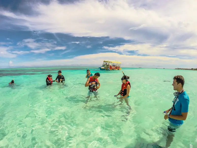 Grupo de turistas em águas cristalinas nas Maldivas preparando-se para mergulho com snorkel. Eles estão vestidos com coletes salva-vidas e equipamentos de mergulho, com um barco colorido ao fundo em um dia ensolarado.