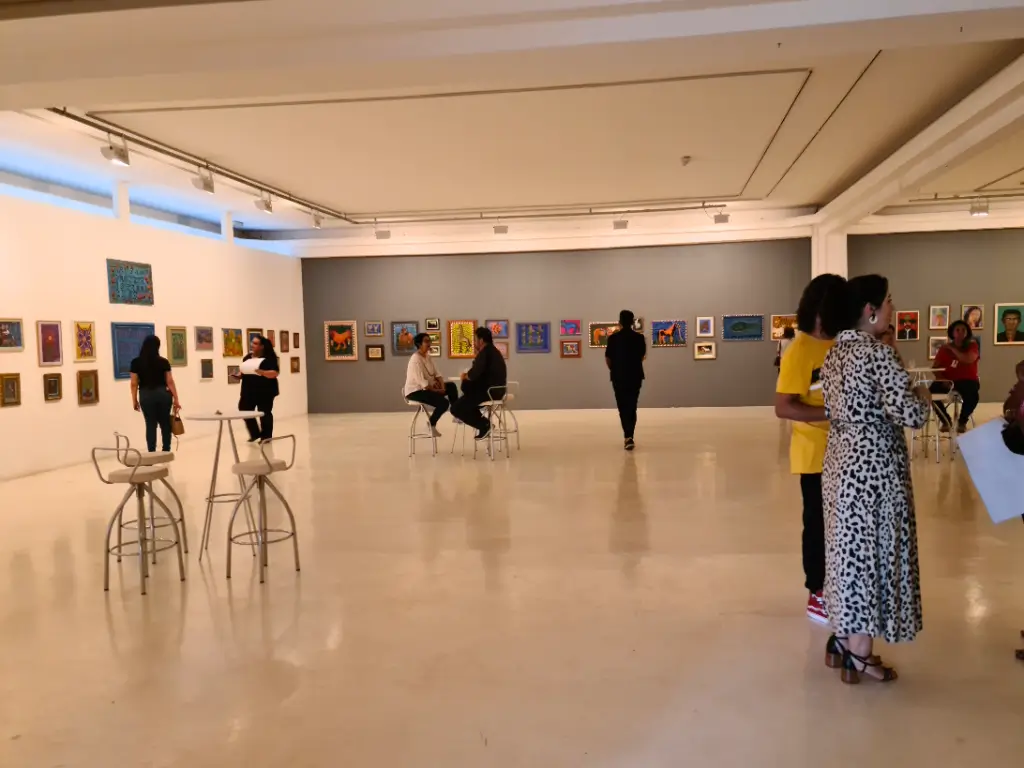 Interior do Museu de Arte Contemporânea de Campinas, com várias pinturas expostas nas paredes e visitantes apreciando as obras e conversando em um ambiente moderno e bem iluminado.