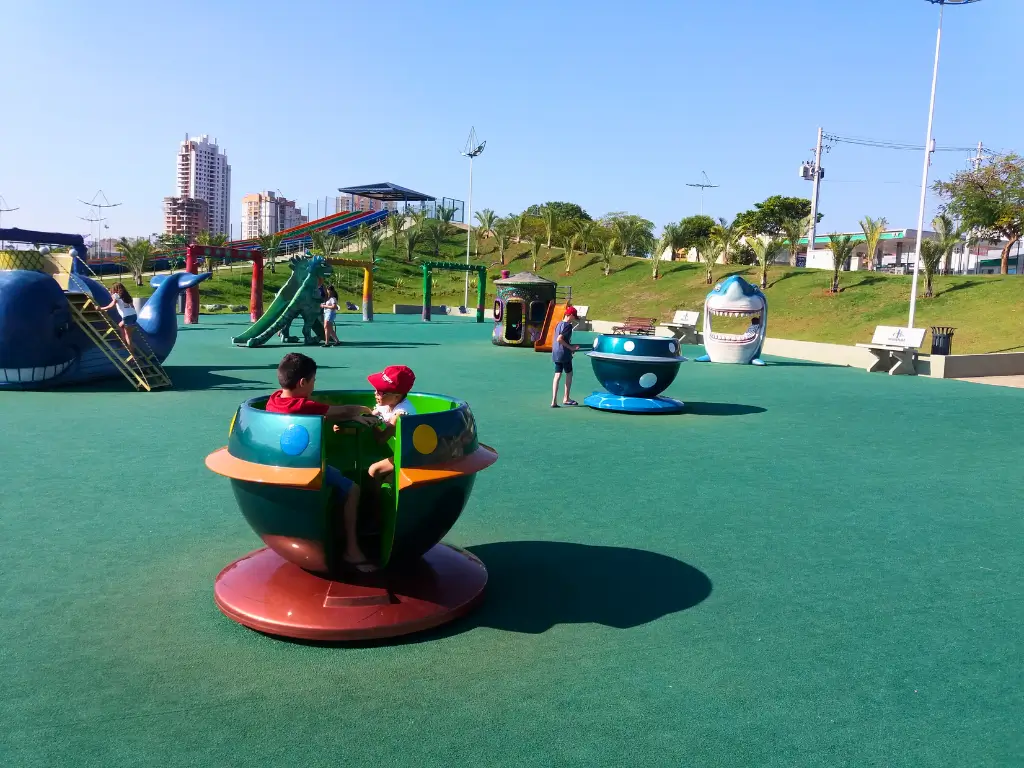 Crianças brincando em um parque infantil colorido com brinquedos temáticos, como um gira-gira e figuras de animais, sob um céu claro, com prédios ao fundo.