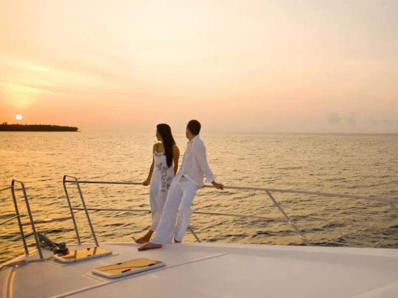 Casal caminhando de mãos dadas na proa de um iate durante o pôr do sol nas Maldivas. Eles estão vestidos de branco, com o oceano ao fundo e um céu laranja ao pôr do sol.