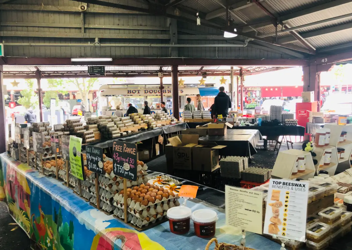 Barracas de ovos e produtos de cera de abelha em um mercado coberto, com clientes ao fundo.