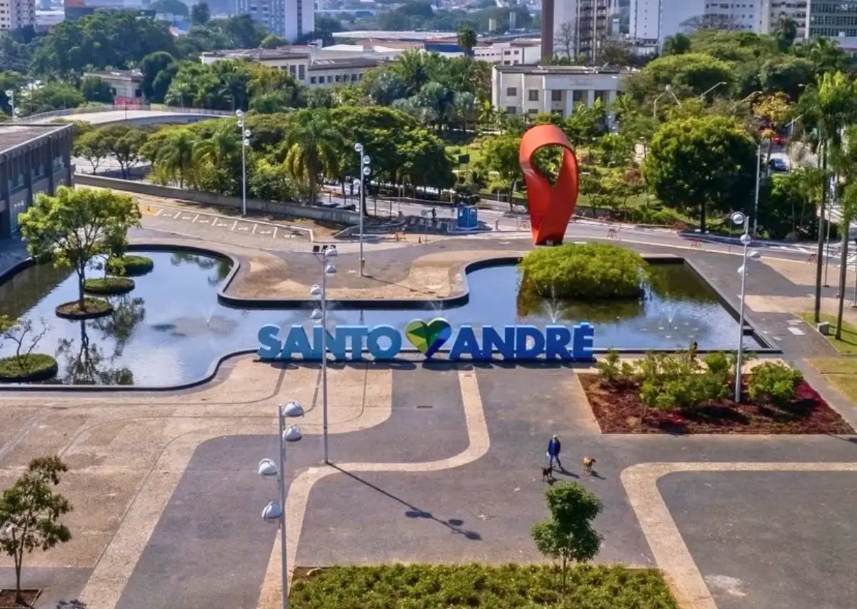Letreiro da entrada de Santo André com o nome da cidade em letras azuis e um coração verde, em frente a um lago artificial e um monumento vermelho, com árvores e edifícios ao fundo.