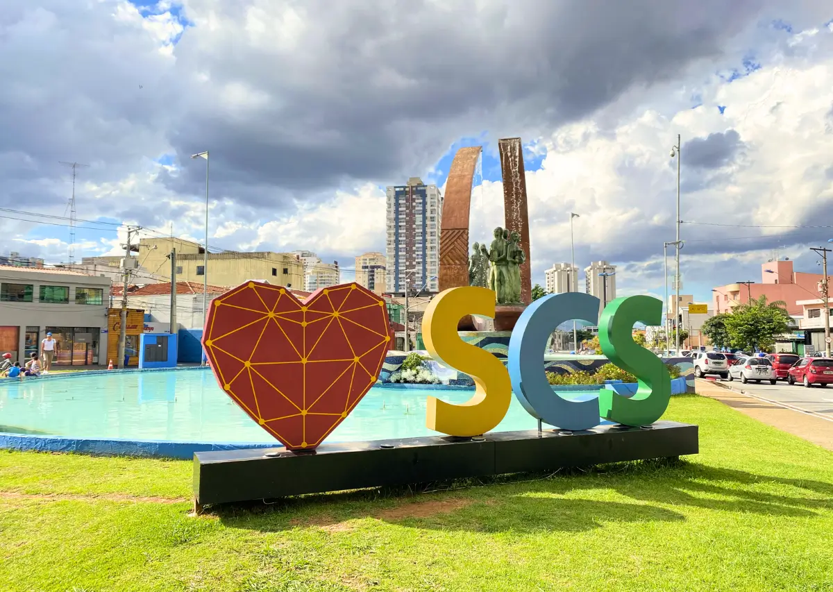 Monumento na entrada de São Caetano do Sul com um grande coração vermelho e as letras 'SCS' em verde, azul e amarelo em frente a uma fonte, com edifícios e um céu nublado ao fundo.