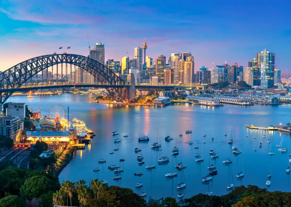 Vista panorâmica noturna do Sydney Harbour Bridge e do horizonte da cidade de Sydney, com barcos ancorados na baía e a Luna Park iluminada em primeiro plano.