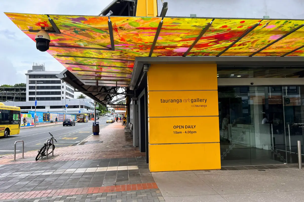 Entrada da Tauranga Art Gallery em um dia nublado, com uma cobertura colorida e vibrante acima da calçada.