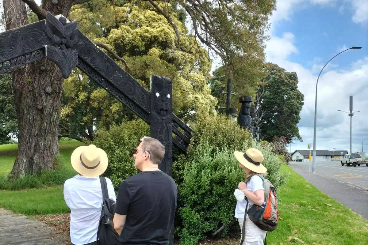 Três turistas examinando uma escultura Maori de madeira ao lado de uma trilha arborizada em Tauranga, Nova Zelândia.