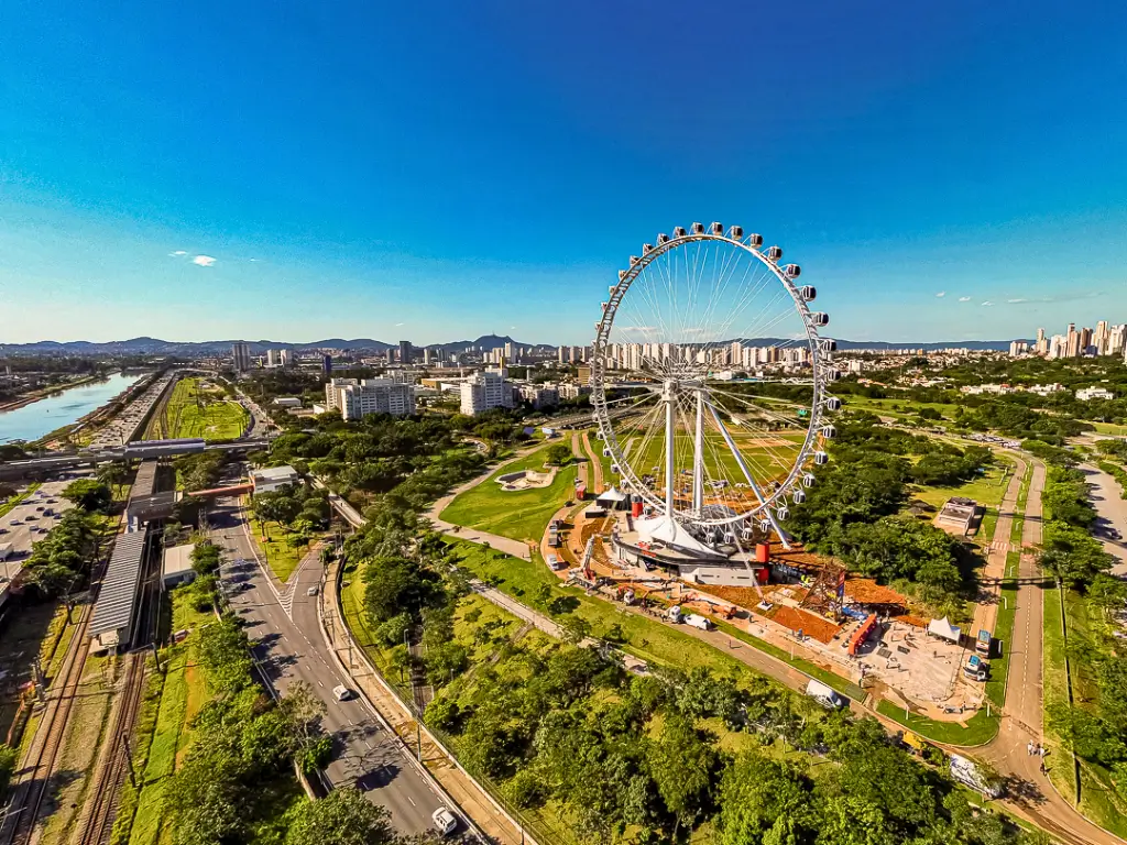 Vista aérea da roda gigante no Parque Villa Lobos, mostrando a cidade de São Paulo ao fundo e o Rio Pinheiros ao lado, em um dia claro e ensolarado.
