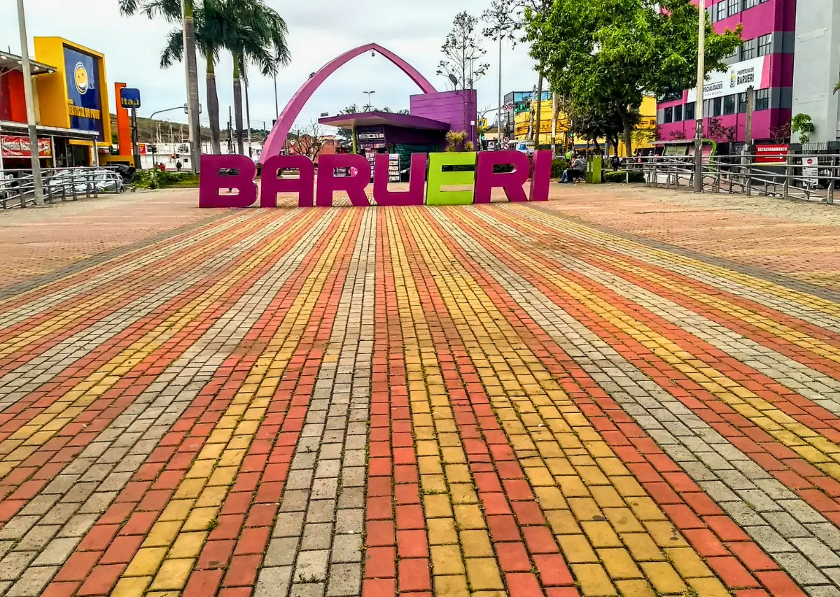 Vista da entrada da cidade de Barueri com um grande letreiro colorido em rosa e branco que lê 'BARUERI'. O chão é pavimentado com tijolos coloridos em tons de amarelo, laranja e vermelho, e ao fundo, árvores e edifícios comerciais modernos.