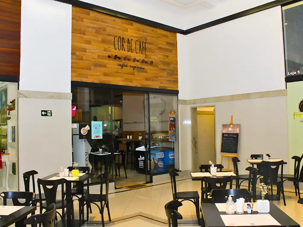 Entrada da cafeteria Cor de Café, com um design moderno que inclui mesas pretas, cadeiras e um letreiro de madeira, localizada em um espaço de shopping com ampla iluminação natural.