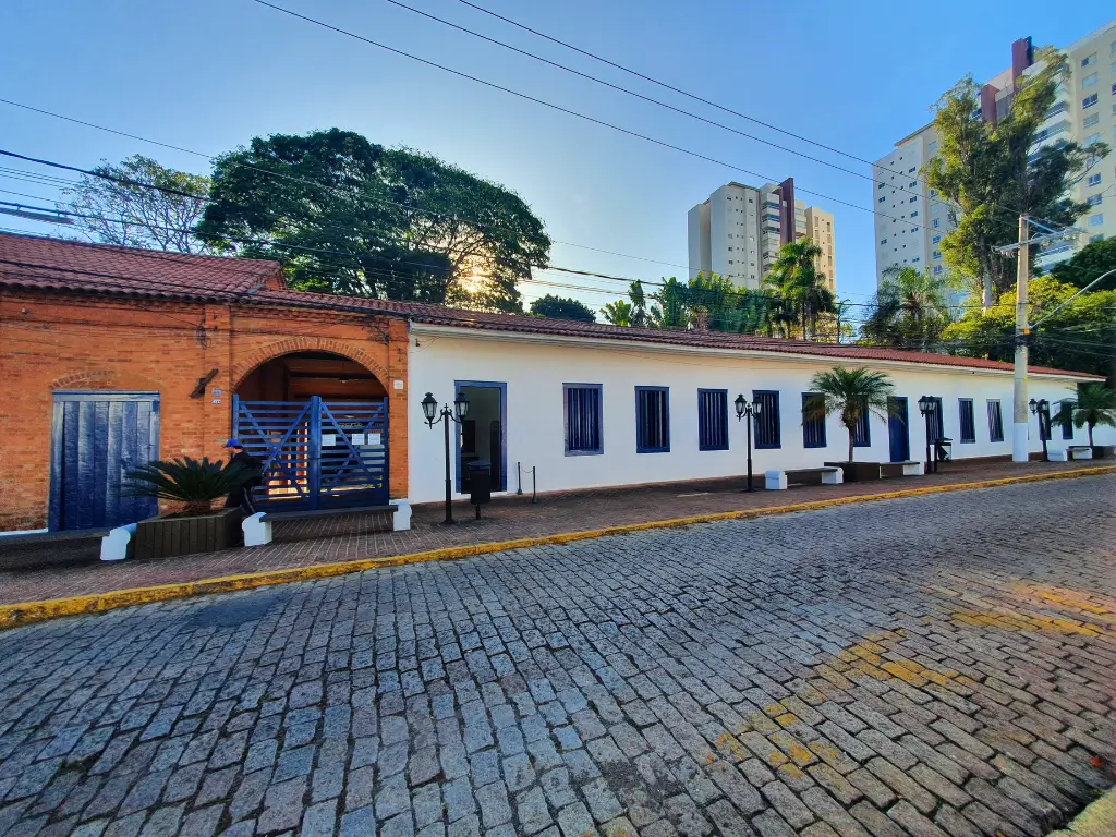 Vista frontal do Casarão Pau Preto em Indaiatuba, um edifício histórico com paredes brancas e janelas azuis, situado em uma rua de paralelepípedos, com prédios modernos ao fundo ao entardecer.