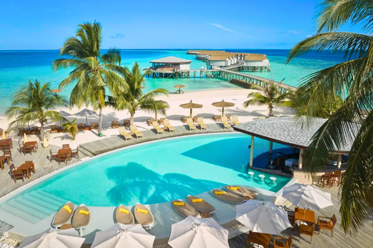 Vista aérea de um resort nas Maldivas com uma grande piscina azul clara, cercada por palmeiras, guarda-sóis brancos e espreguiçadeiras de madeira. Ao fundo, um píer estende-se sobre o oceano turquesa com mais cabanas e um bar sobre a água.