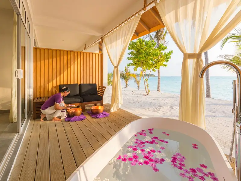 Resort de praia nas Maldivas com um terraço privado. Uma mulher em uniforme roxo prepara uma banheira de hidromassagem com pétalas de flores cor-de-rosa. Ao lado, um espaço de lounge ao ar livre com sofá, mesa e vista para palmeiras e o mar ao fundo.