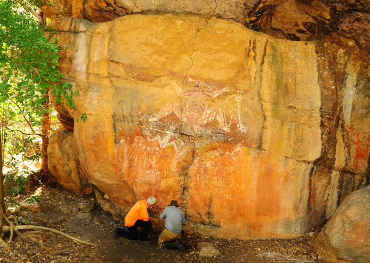 Duas pessoas observam pinturas rupestres aborígenes em uma rocha grande, cercada por vegetação.