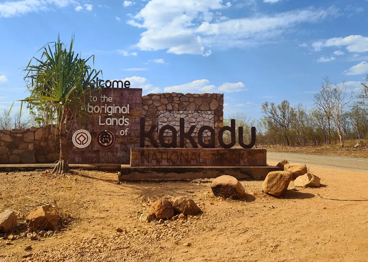 Pontos Turísticos da Austrália: Entrada do Parque Nacional de Kakadu, com um letreiro de boas-vindas nas terras aborígenes, cercado por uma paisagem árida e céu azul.