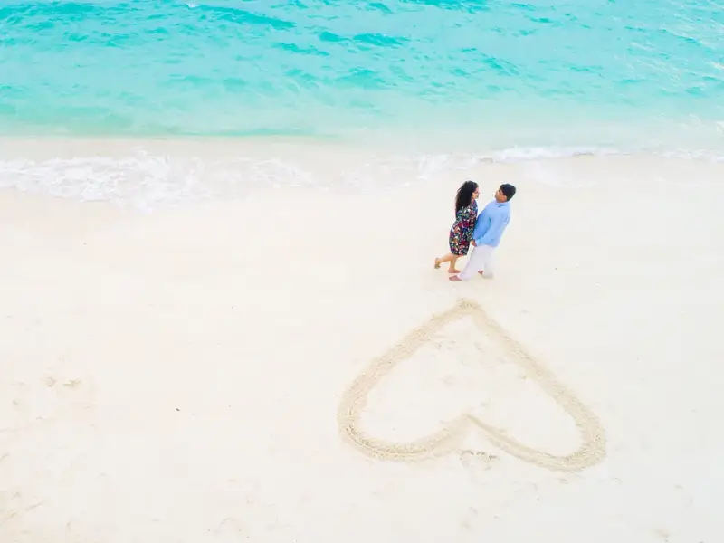 Casal de mãos dadas na praia com coração desenhado na areia ao fundo