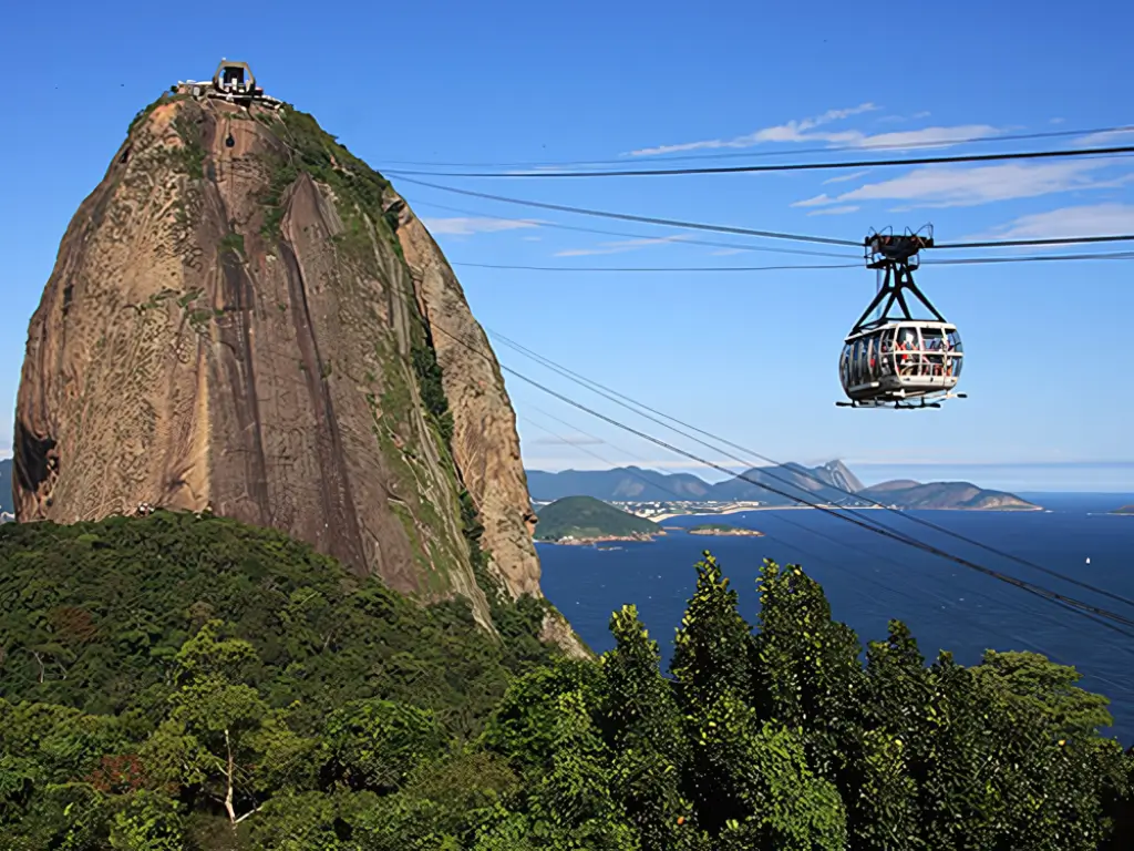 Foto mostrando o bondinho do Pão de Açúcar em movimento, com o imponente morro e o oceano Atlântico ao fundo, em um dia claro.