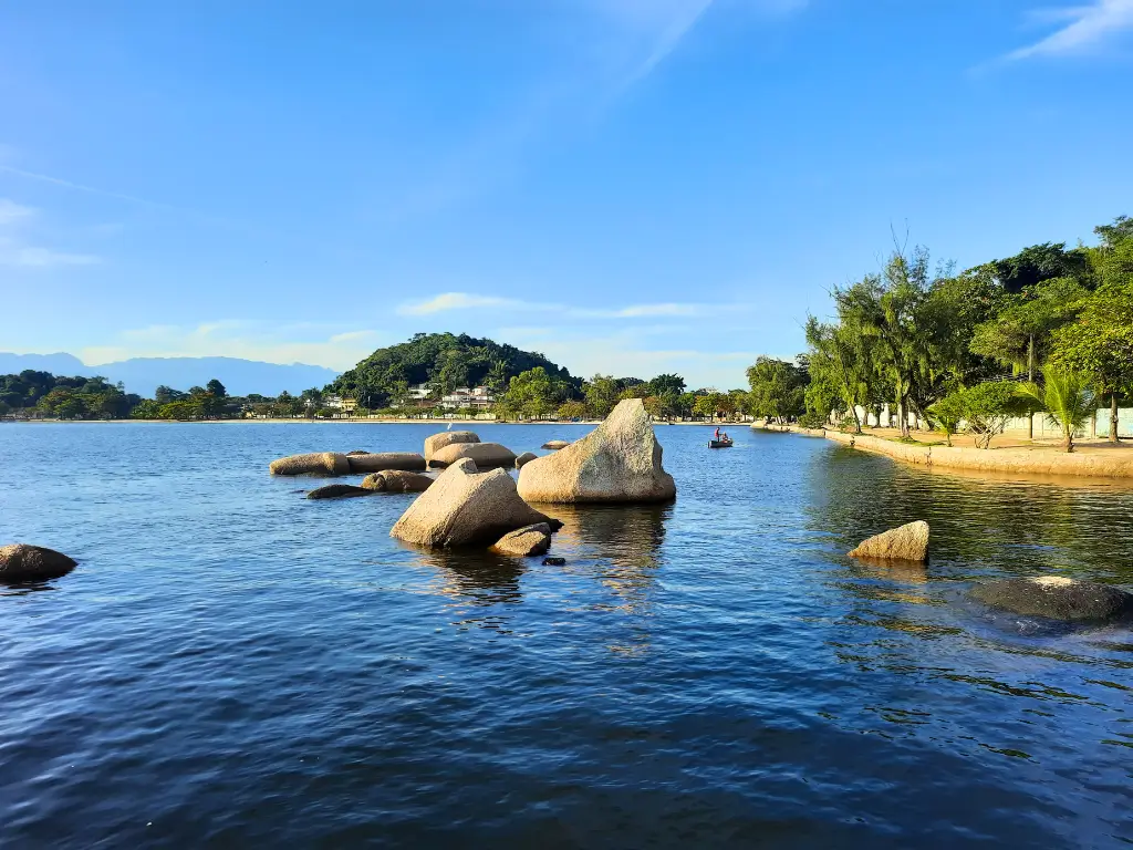 Ilha de Paquetá, vista serena de um lago com rochas grandes à beira d'água, árvores ao longo da margem e montanhas ao fundo em um dia ensolarado.