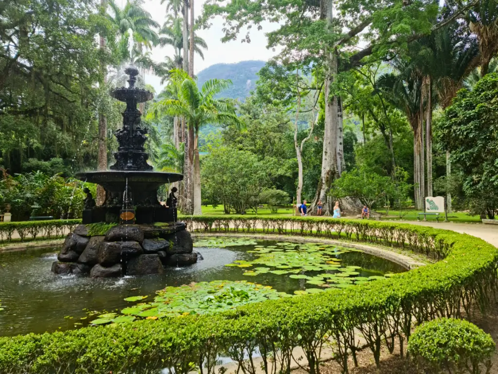 Foto de uma fonte ornamental no Jardim Botânico do Rio de Janeiro, cercada por um pequeno lago com lírios d'água, emoldurada por árvores altas e exuberantes jardins.