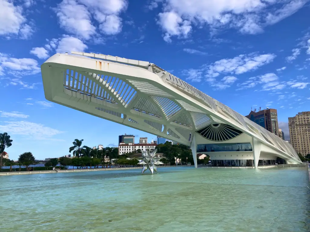 Foto do Museu do Amanhã no Rio de Janeiro, uma estrutura moderna e futurista localizada à beira de um lago artificial, sob um céu azul com nuvens dispersas.