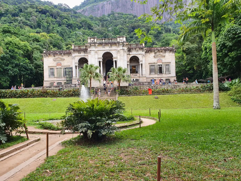 Vista do Parque Lage no Rio de Janeiro, mostrando um edifício histórico com arquitetura clássica, cercado por jardins verdes, uma fonte ativa e turistas caminhando no local.