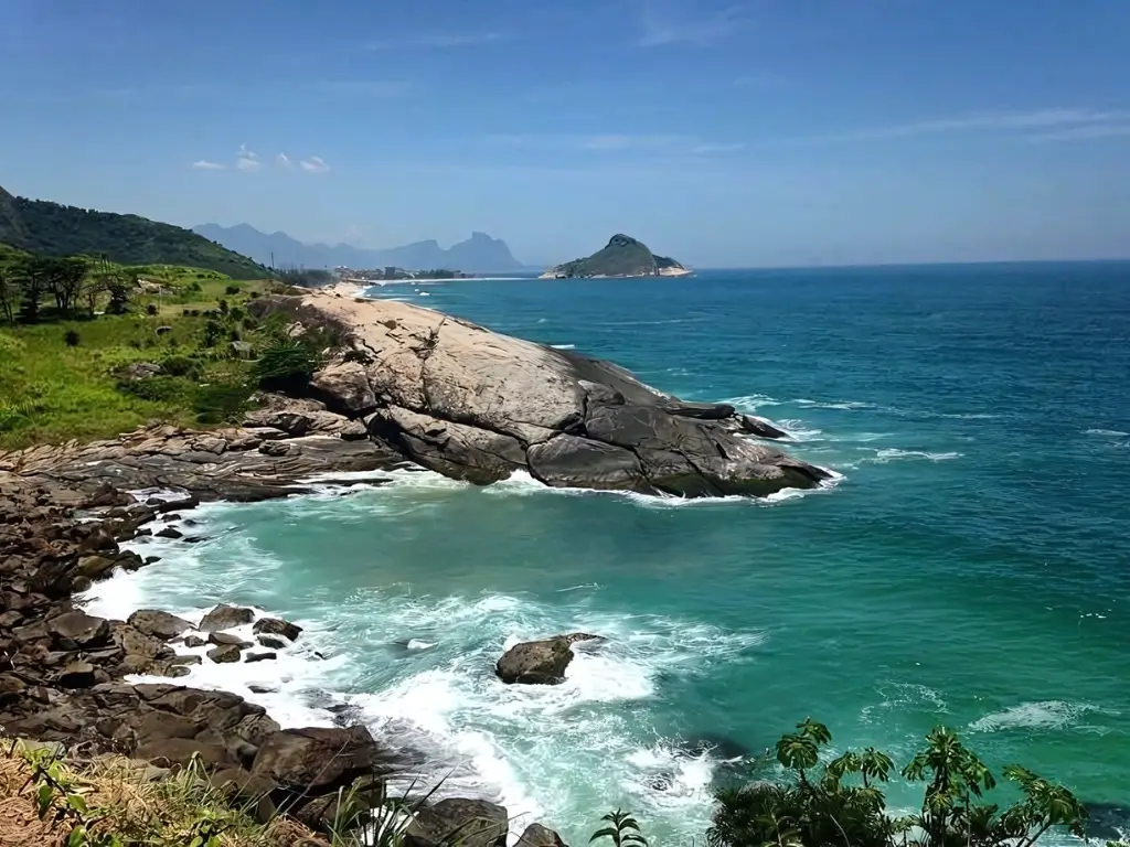 Praia de Grumari, vista panorâmica de uma costa rochosa no Rio de Janeiro com águas turquesa batendo contra as rochas, uma praia ao fundo e montanhas distantes sob um céu claro.