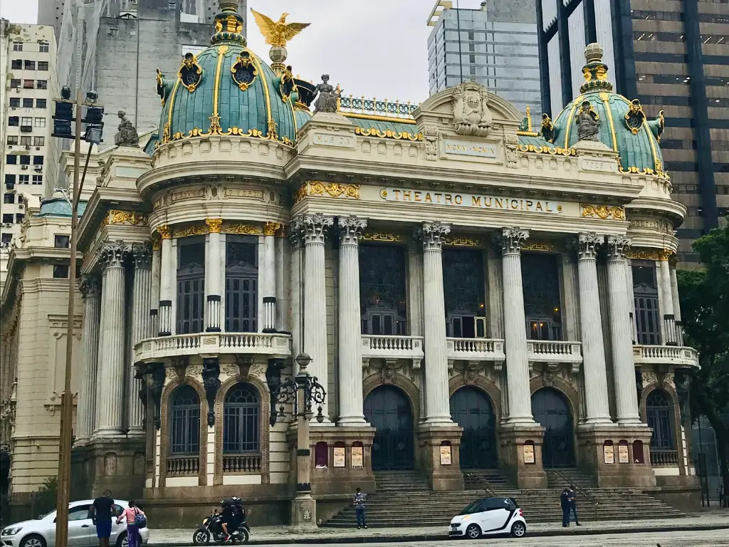 Foto do Theatro Municipal do Rio de Janeiro, mostrando sua fachada neoclássica ornamentada com detalhes dourados e estátuas, localizado em uma praça pavimentada com calçadas de pedra, em um dia nublado.