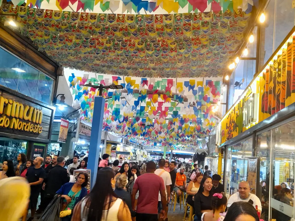 Feira de São Cristóvão, imagem vibrante de um mercado coberto decorado com bandeirinhas coloridas durante uma festa, mostrando muitas pessoas andando entre barracas e outras sentadas em um café, desfrutando da atmosfera festiva.